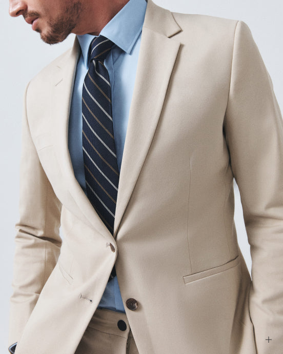A close-up of a man wearing a light beige suit jacket, a light blue dress shirt, and a navy blue striped tie, shown from the chin down against a plain light gray background.